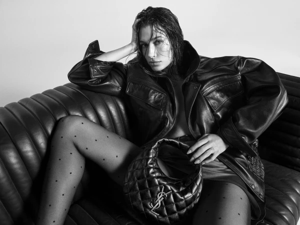 Hailey Bieber wearing an oversized leather jacket seated on a leather sofa holding a quilted handbag in a black and white fashion campaign image