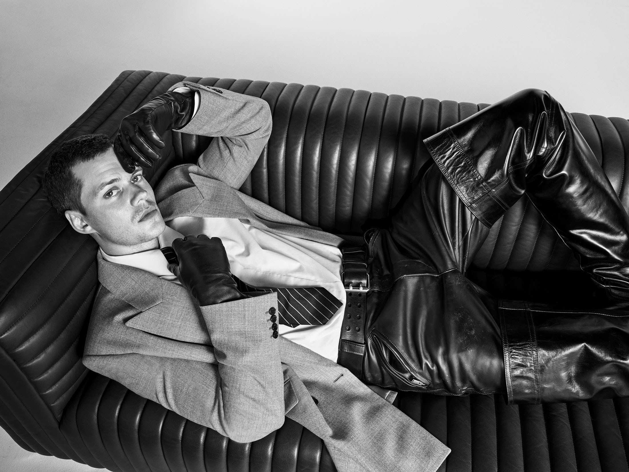 Man wearing a grey suit jacket and black leather trousers reclining on a ridged leather sofa in a black and white fashion portrait.