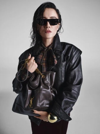 Model wearing dark sunglasses, black leather jacket and carrying a brown leather shoulder bag against a grey seamless background