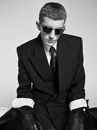 Model wearing dark sunglasses, a black double-breasted suit, white cuffs, and leather gloves against a plain light background