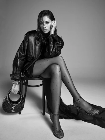 Woman in dark leather jacket and polka dot tights seated on a chair holding a quilted handbag against a plain grey background