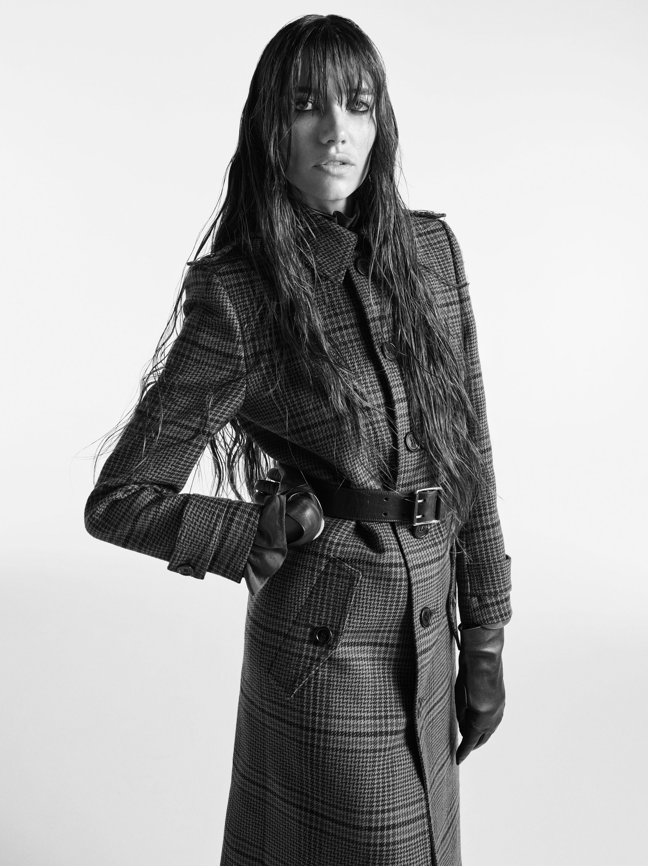 Model in dark plaid coat with belt and leather gloves posing against a plain background