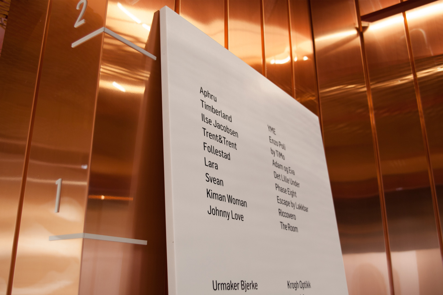 White directory sign mounted on polished copper elevator wall with brand names