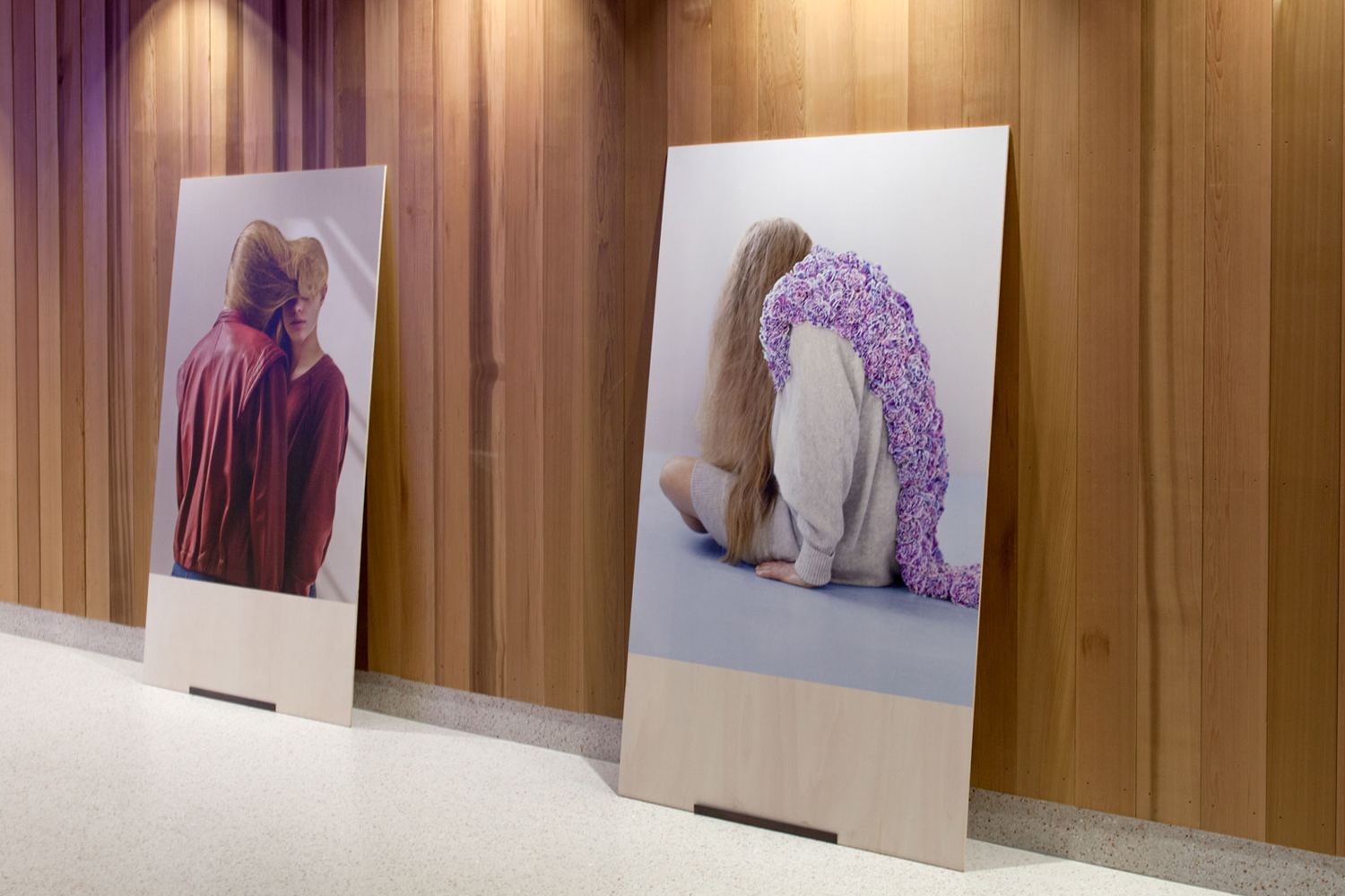 Two large vertical posters featuring fashion photography displayed against a vertical wooden slat wall on a terrazzo floor.