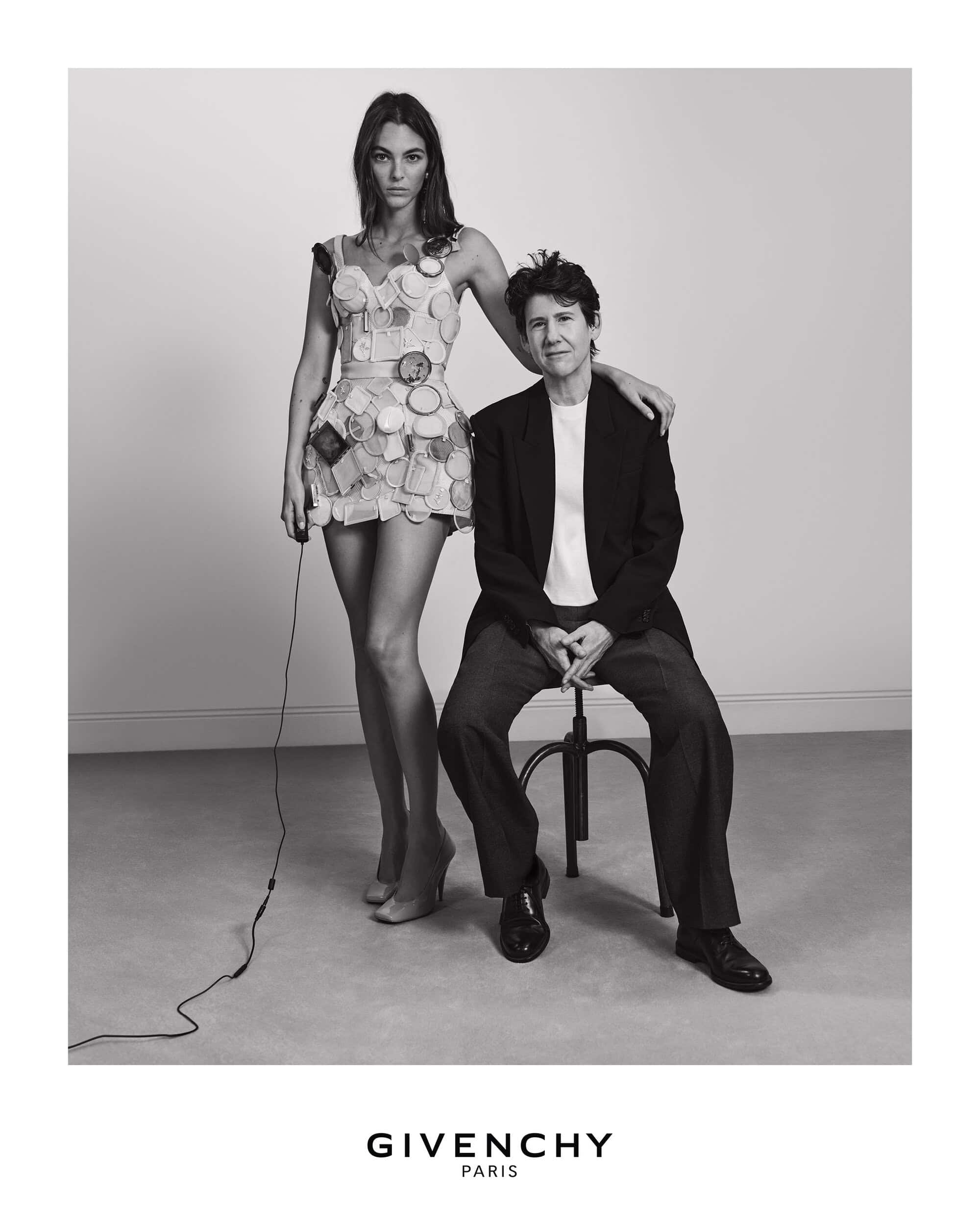 Model standing next to seated designer in black blazer, wearing a dress adorned with small mirrors against a white backdrop.