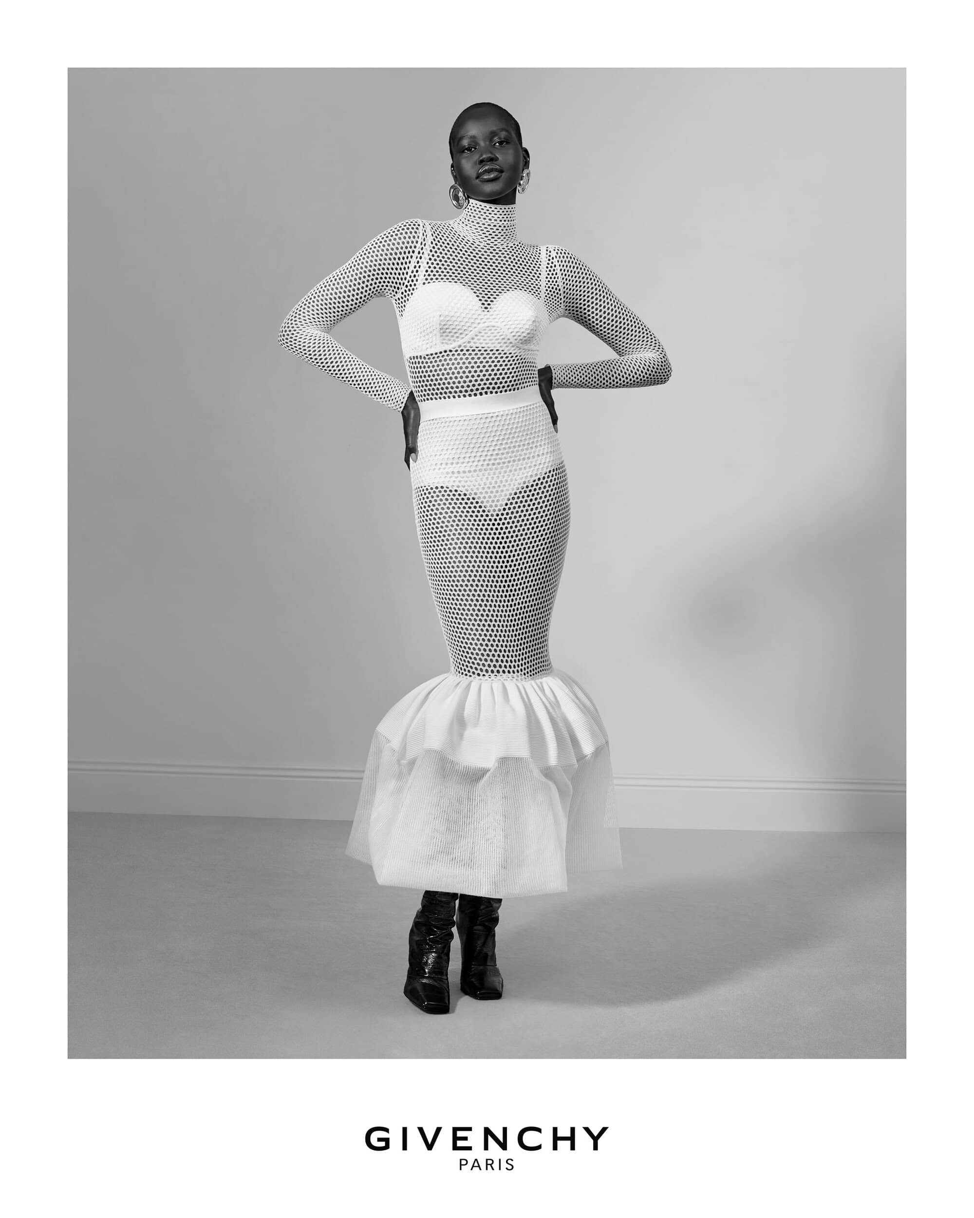 An adult female model poses in a sheer white knitted dress with a ruffled hem and black boots against a plain backdrop.