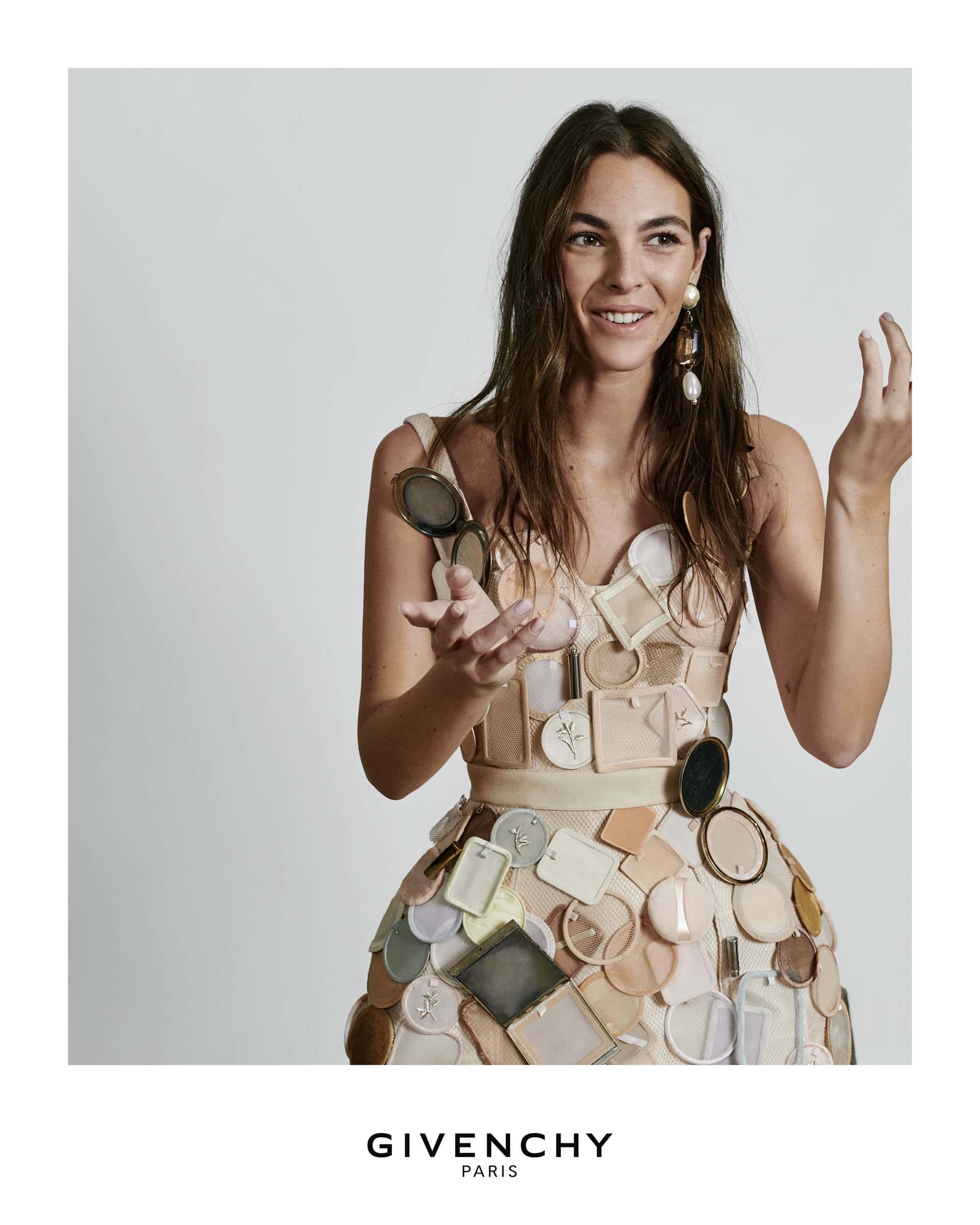 Model wearing a structured dress adorned with cosmetic compacts and mirrors against a light grey seamless background