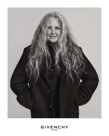 Portrait of an older woman with long grey hair wearing a dark coat against a plain background for Givenchy Fall 2025.