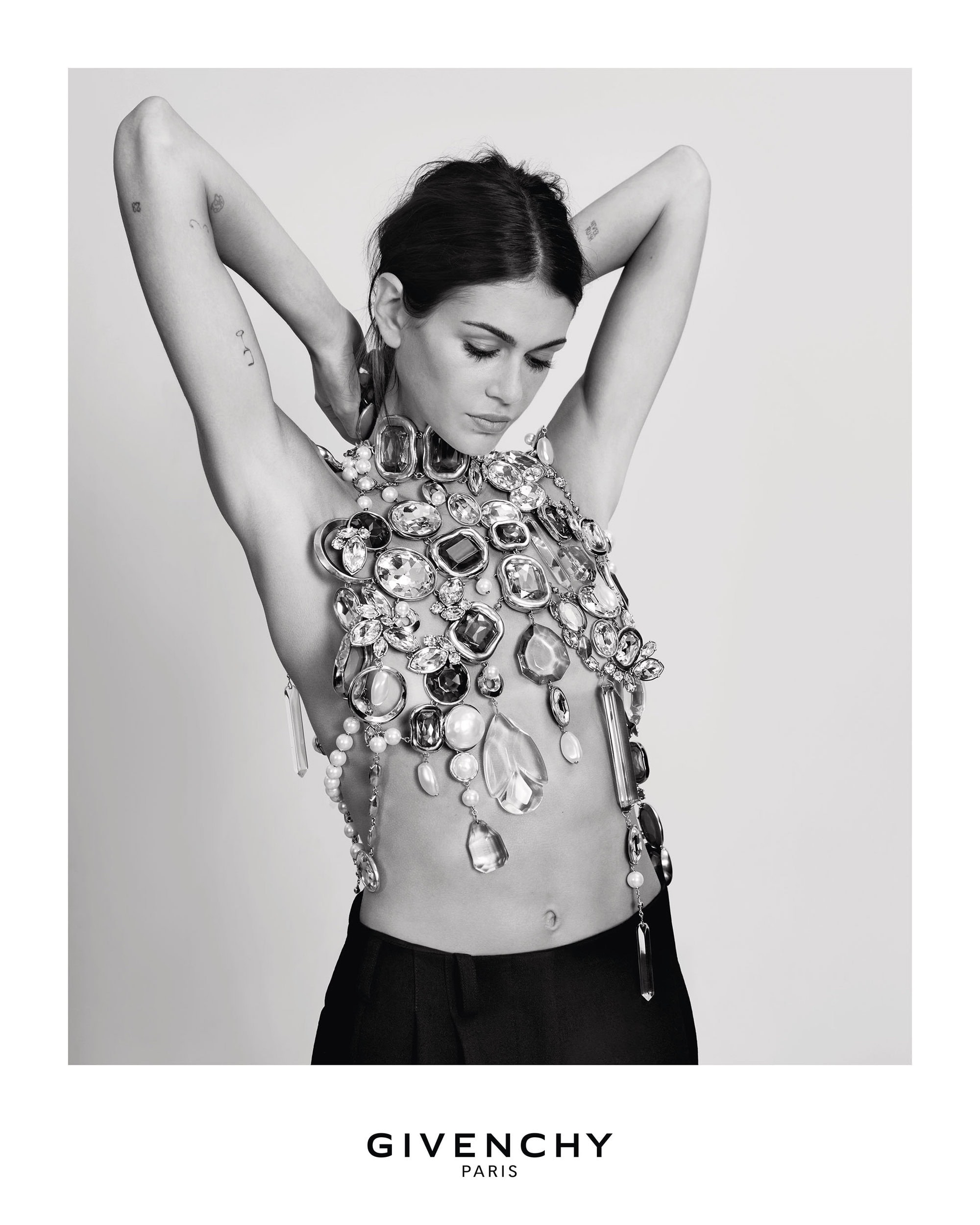 Model Kaia Gerber wearing an elaborate crystal-embellished halter top and dark trousers against a white backdrop for Givenchy.
