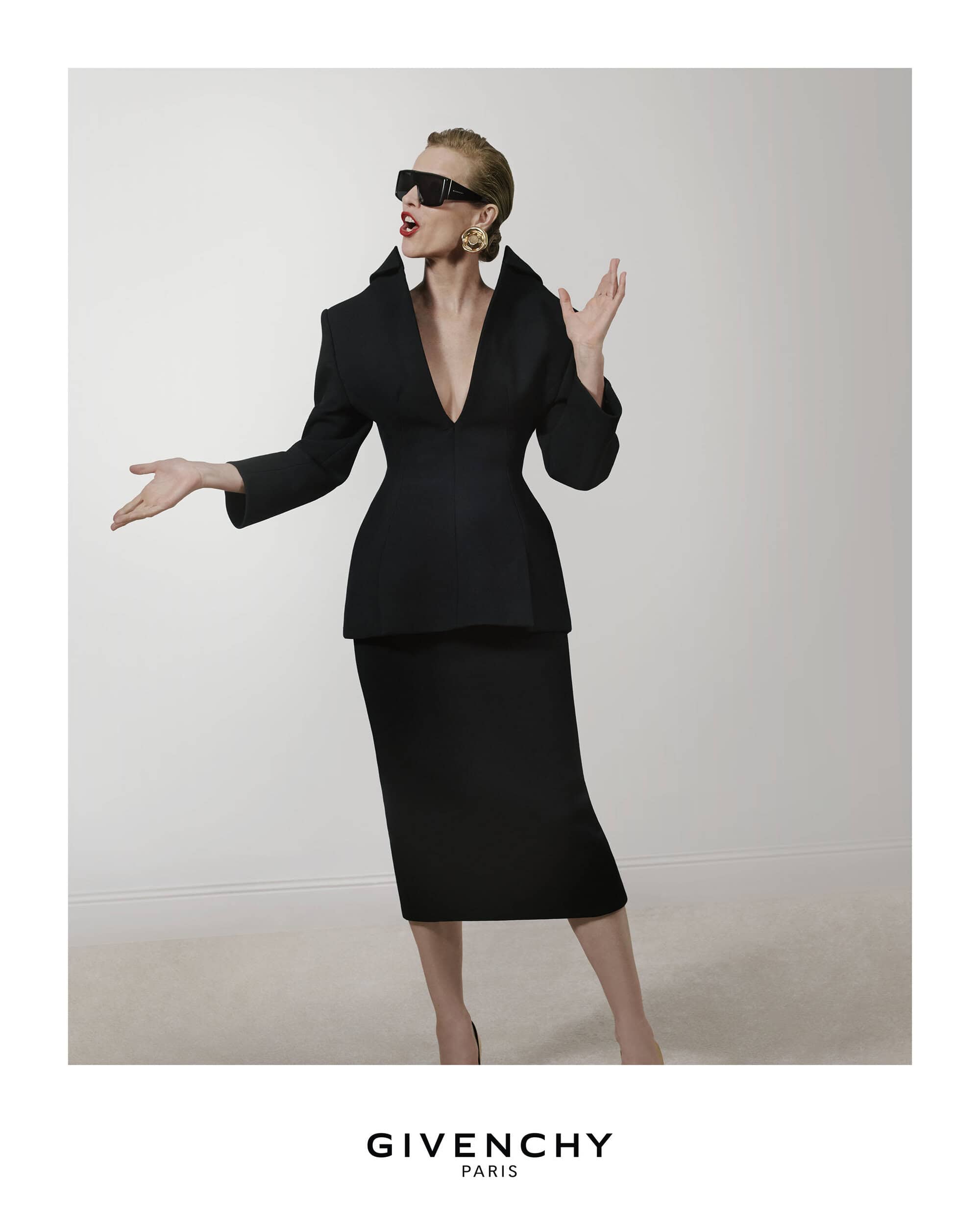 Model Eva Herzigova wearing black blazer and matching skirt with dark sunglasses and gold earring against white backdrop