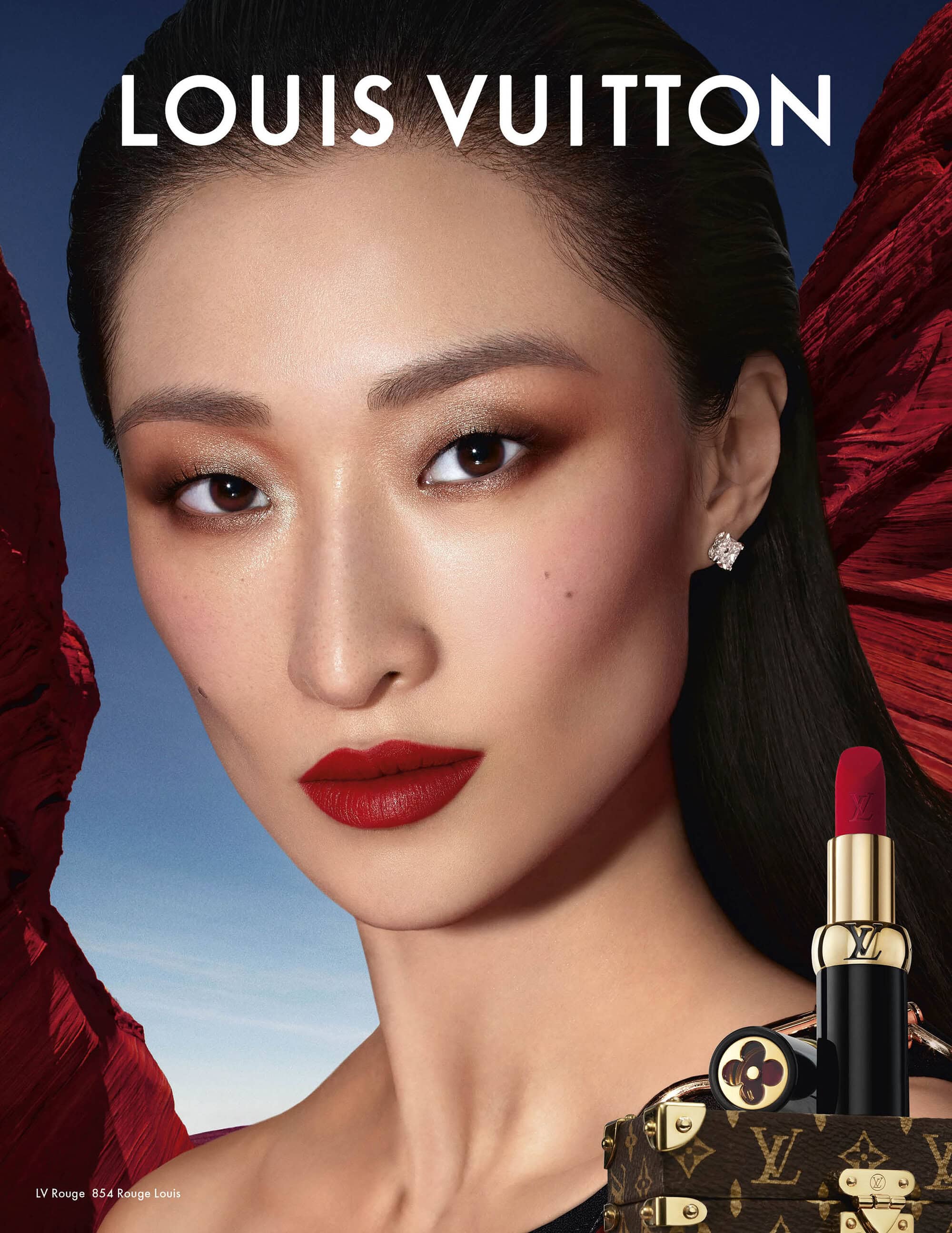 Close-up beauty portrait of an East Asian model wearing bold red lipstick and a gold earring against a textured red background.