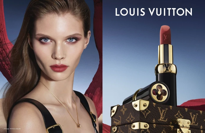 Close-up beauty portrait of a model next to a Louis Vuitton lipstick standing atop a miniature monogram trunk against a blue sky.