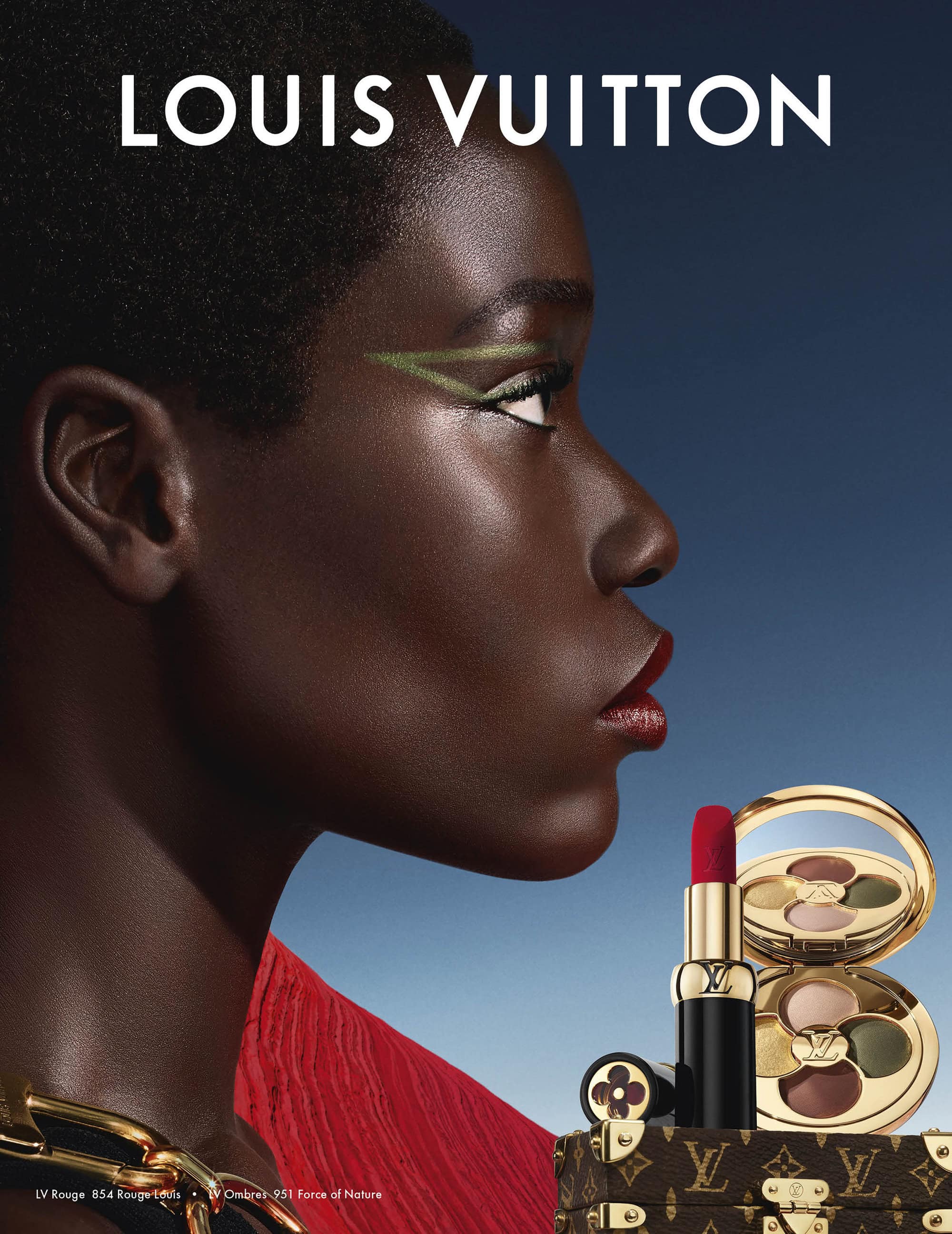 Close-up profile of a black woman with red lipstick and green graphic eyeliner next to gold makeup products and a miniature trunk.