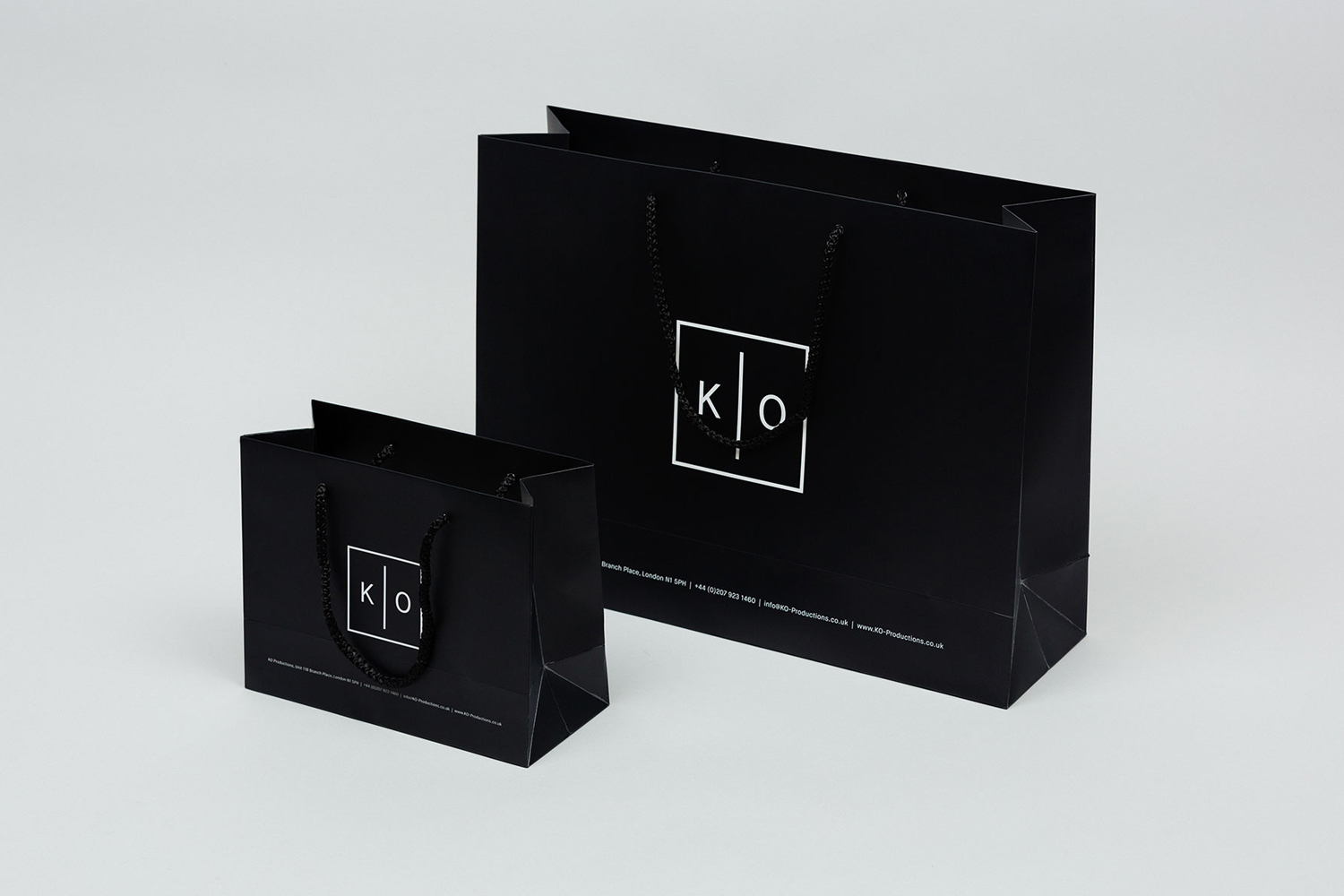 Two black paper shopping bags with white square logo branding and dark rope handles on a white surface.