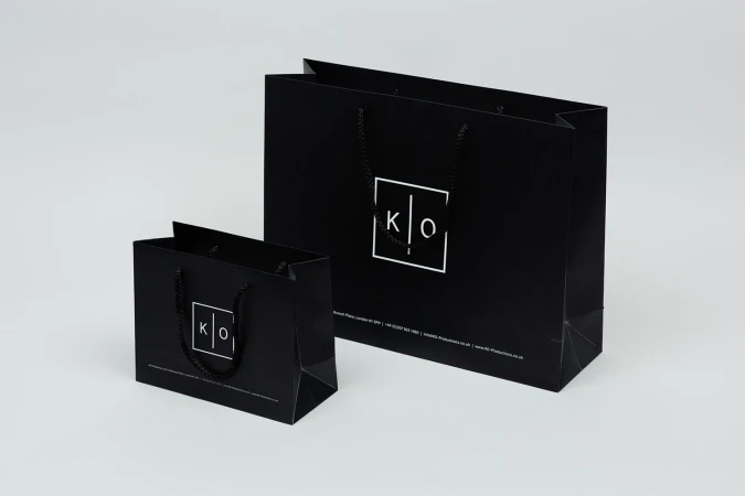 Two black paper shopping bags with white square logo branding and dark rope handles on a white surface.