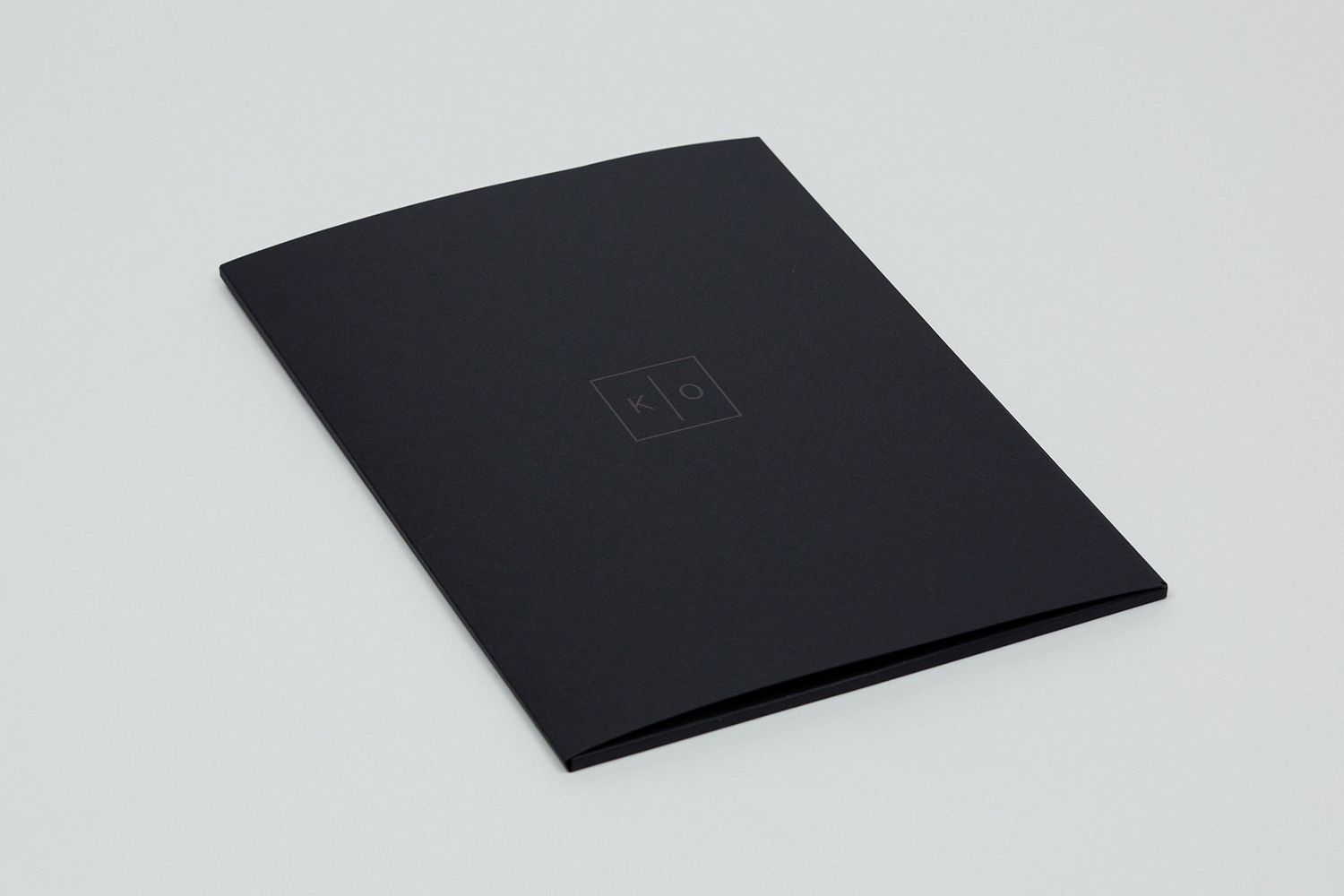 A sleek black booklet with a minimalist KO Productions logo rests centrally on a clean neutral grey surface.