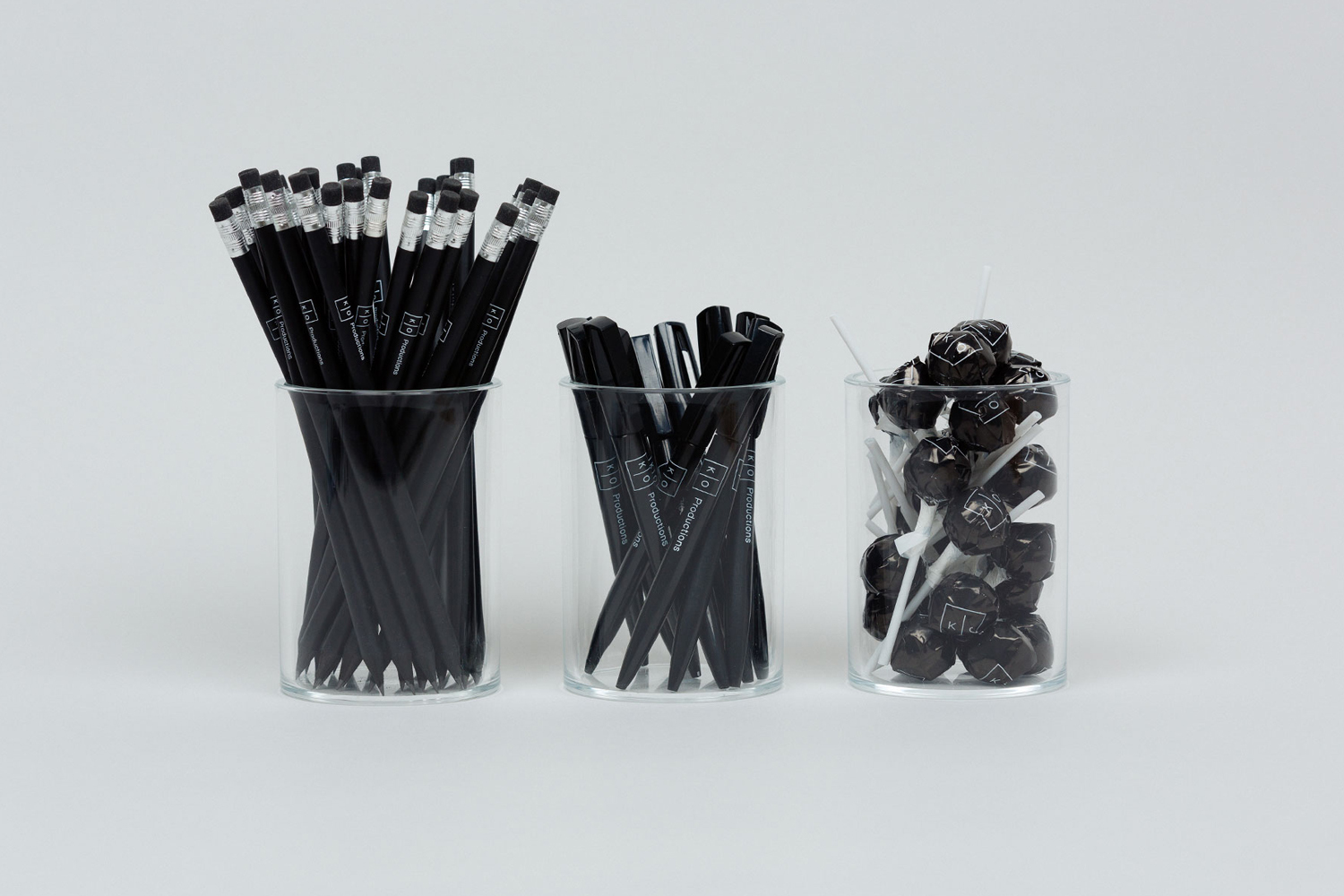 Three clear glass jars containing black pencils, black pens, and black lollipops against a plain white background.