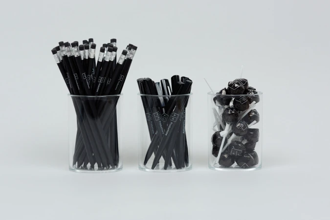 Three clear glass jars containing black pencils, black pens, and black lollipops against a plain white background.
