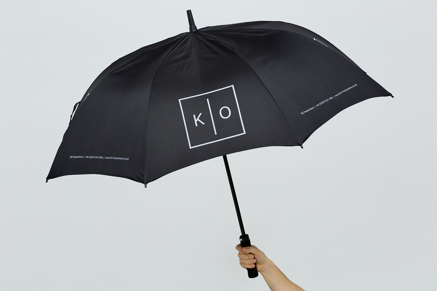 Hand holding a black umbrella with a white box logo featuring letters K and O against a seamless grey background