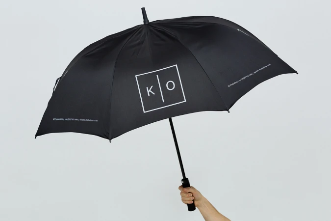 Hand holding a black umbrella with a white box logo featuring letters K and O against a seamless grey background