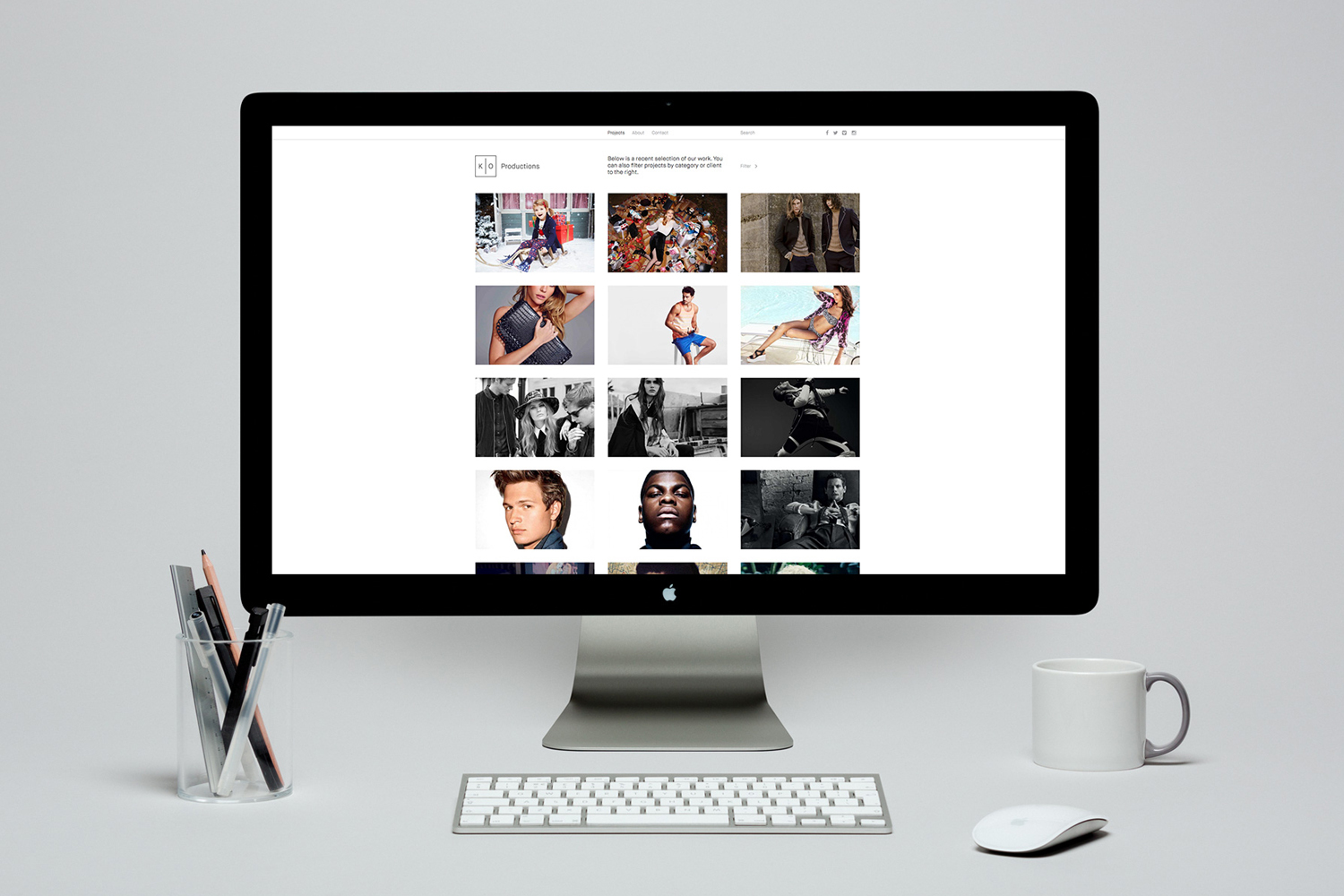 Desktop computer displaying a creative portfolio website with a keyboard, mouse, coffee mug, and a cup of pens on a white desk