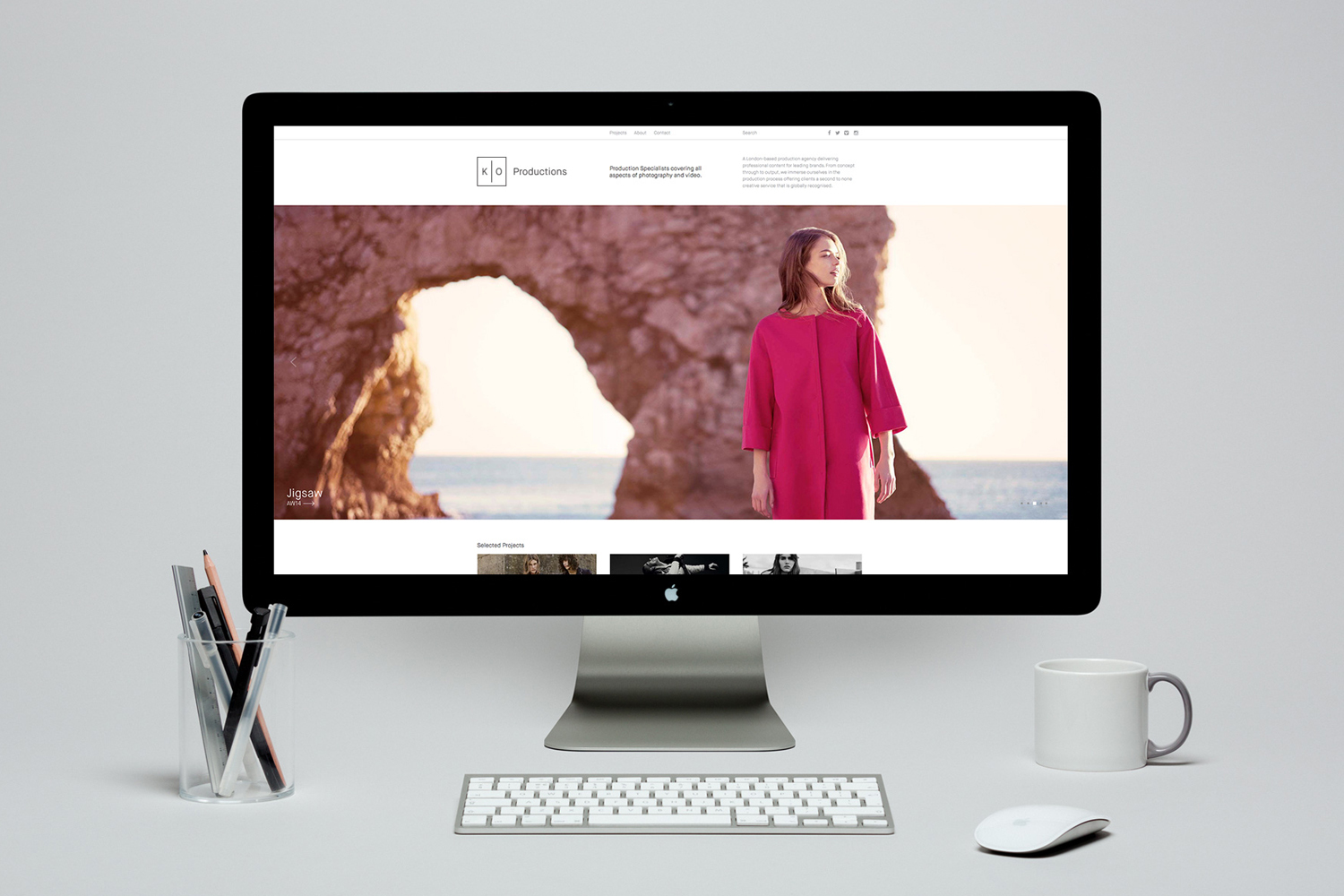 Computer monitor displaying KO Productions website mockup with a pink coat editorial image against a rocky coastal background.