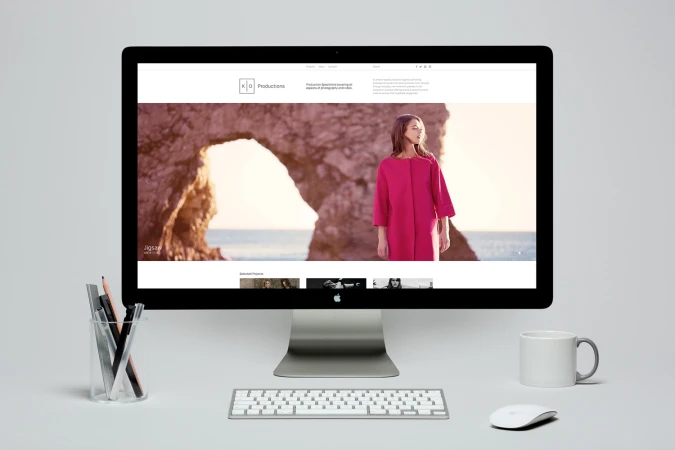 Computer monitor displaying KO Productions website mockup with a pink coat editorial image against a rocky coastal background.