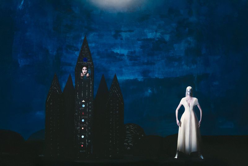 Two women against a deep blue background, one standing in a light gown and one positioned within a dark, ornate, castle-like structure.