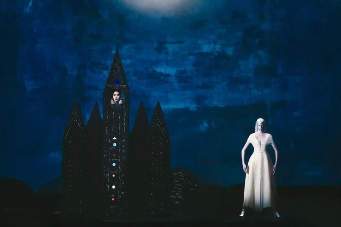 Two women against a deep blue background, one standing in a light gown and one positioned within a dark, ornate, castle-like structure.