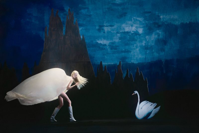 A woman in a flowing white cape and silver boots stands near a swan against a painted backdrop of a blue sky and castle.