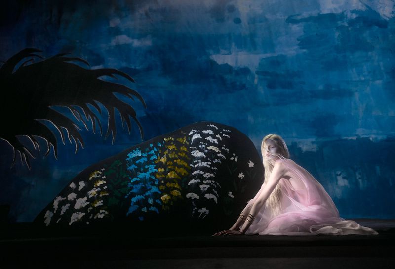 Adult woman with long blonde hair, wearing a pink dress sitting with hands outstretched on a dark surface in front of a blue painted backdrop.