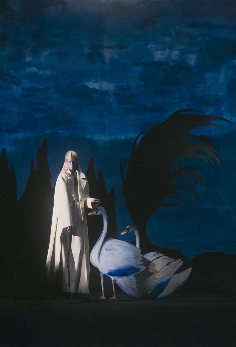 Blond-haired person in white coat poses with large swan props against a blue and black painted backdrop in a fashion style photo.