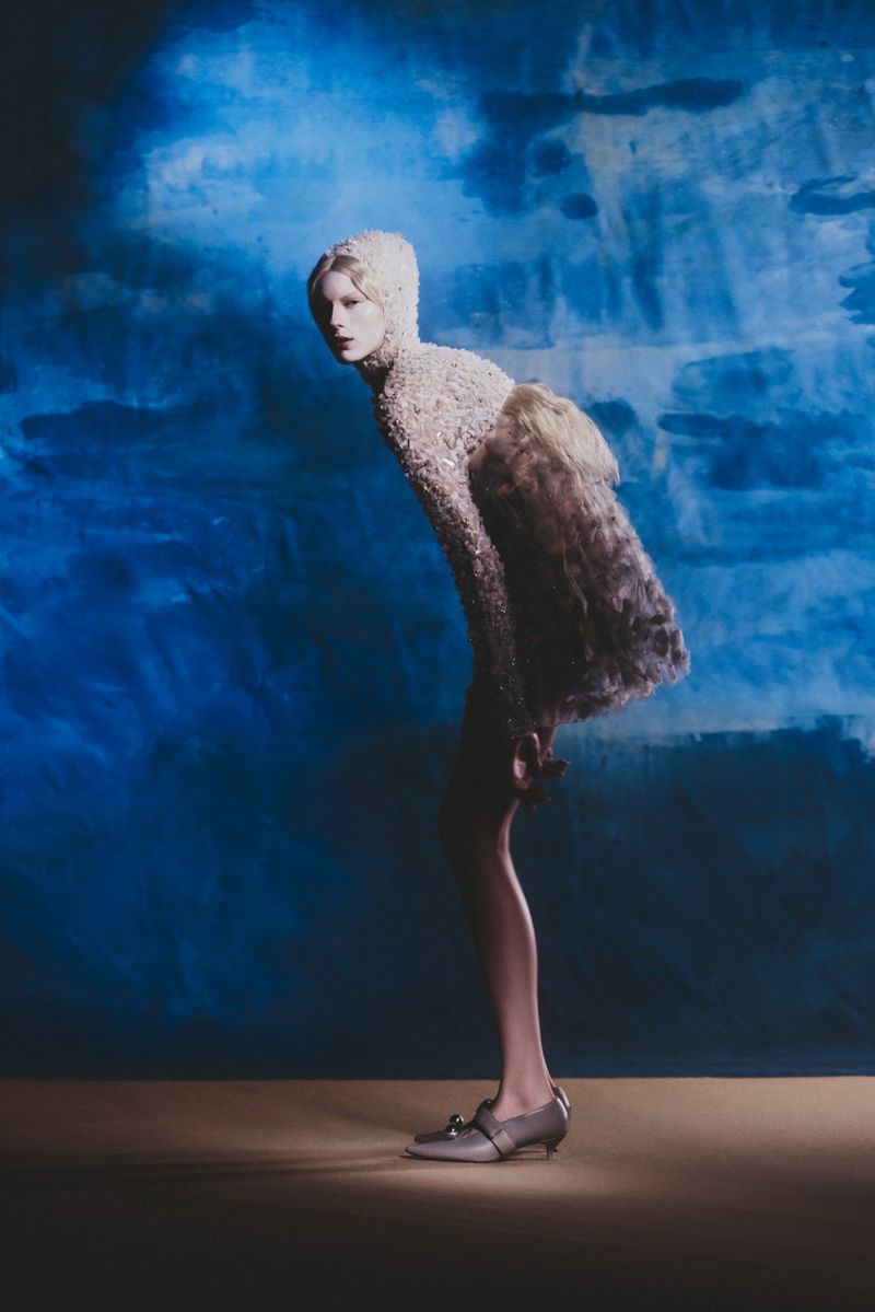 Elegant fashion image of a young woman posing in a beige embellished dress with a fur detail against a blue mottled background in studio light.