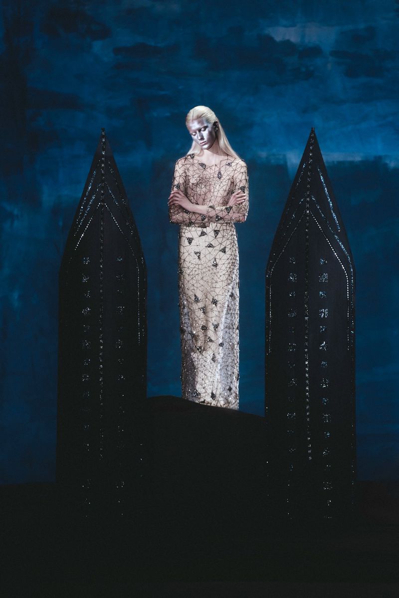 Fashion shot of a slender woman with face paint in a long gown standing between two dark, spired objects against a textured blue background.