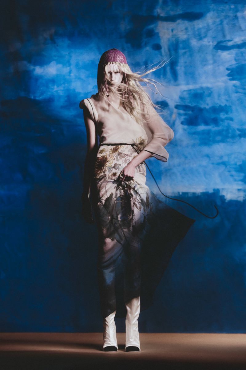 Full-length fashion shot of a blonde female model wearing a light brown top, floral skirt, and white boots against a blue background.