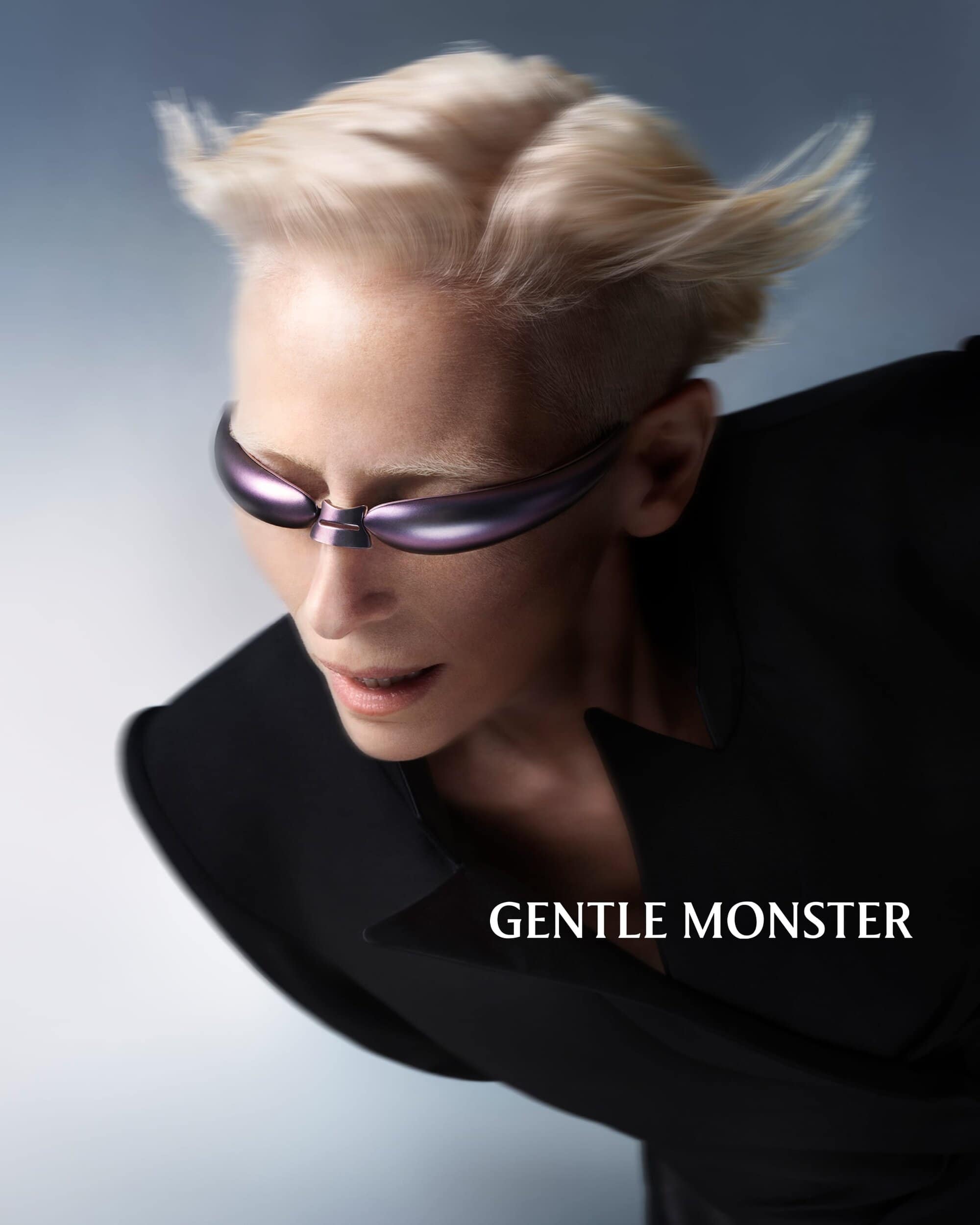 Tilda Swinton wearing metallic purple futuristic sunglasses and a black jacket against a grey gradient background