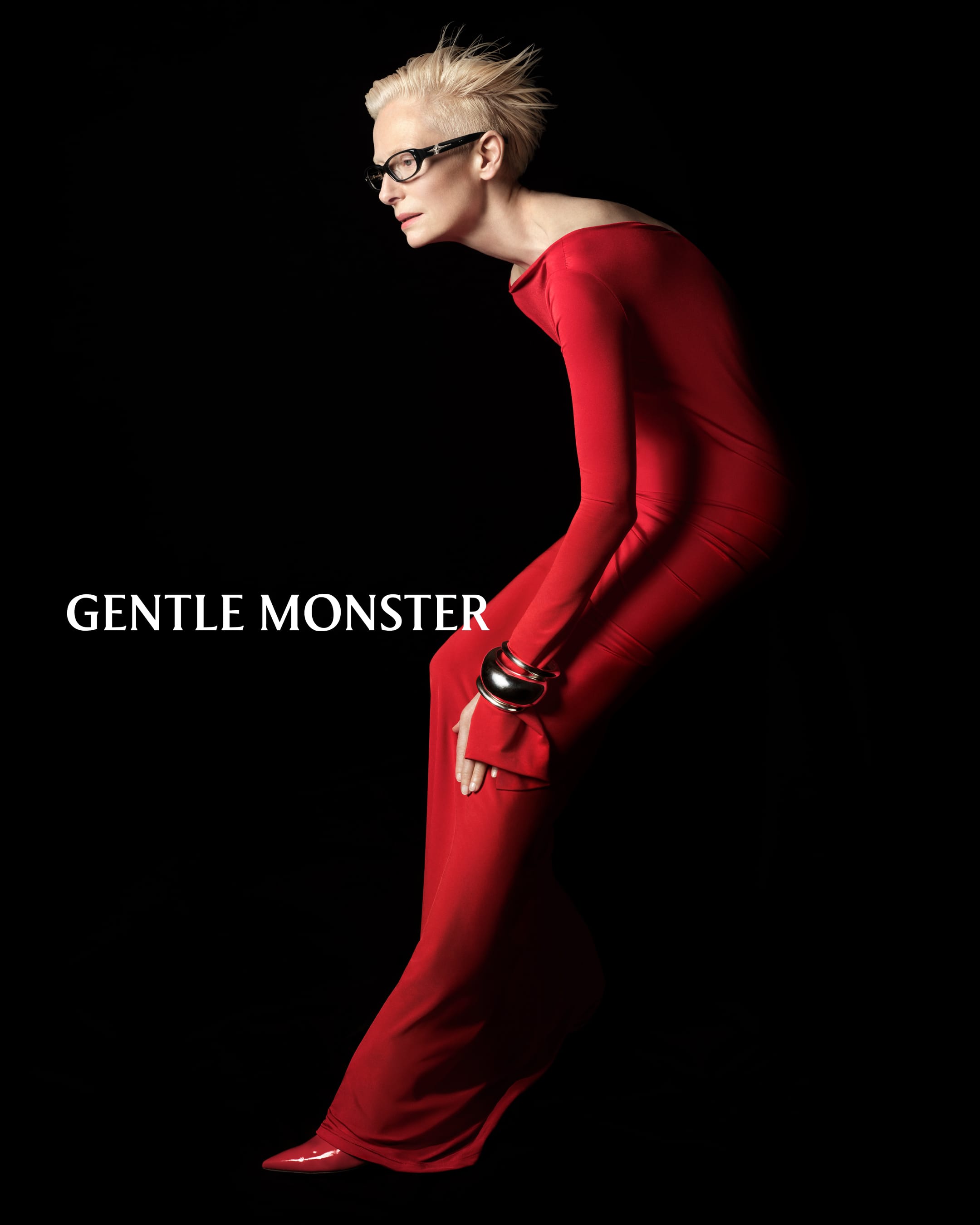 Tilda Swinton in a long red dress wearing dark eyeglasses against a black background for Gentle Monster