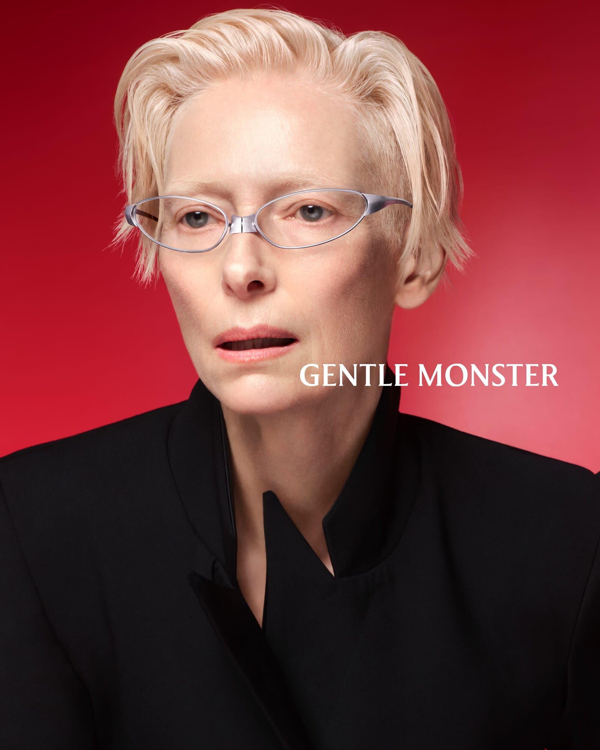 Tilda Swinton wearing metallic eyeglasses and a black jacket against a red background for Gentle Monster.
