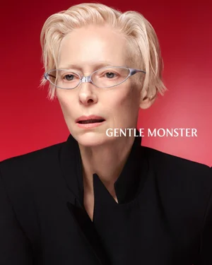 Tilda Swinton wearing metallic eyeglasses and a black jacket against a red background for Gentle Monster.