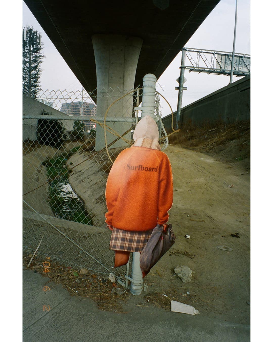 Person wearing an orange sweatshirt with lettering and a plaid skirt standing under an overpass near a chain-link fence.