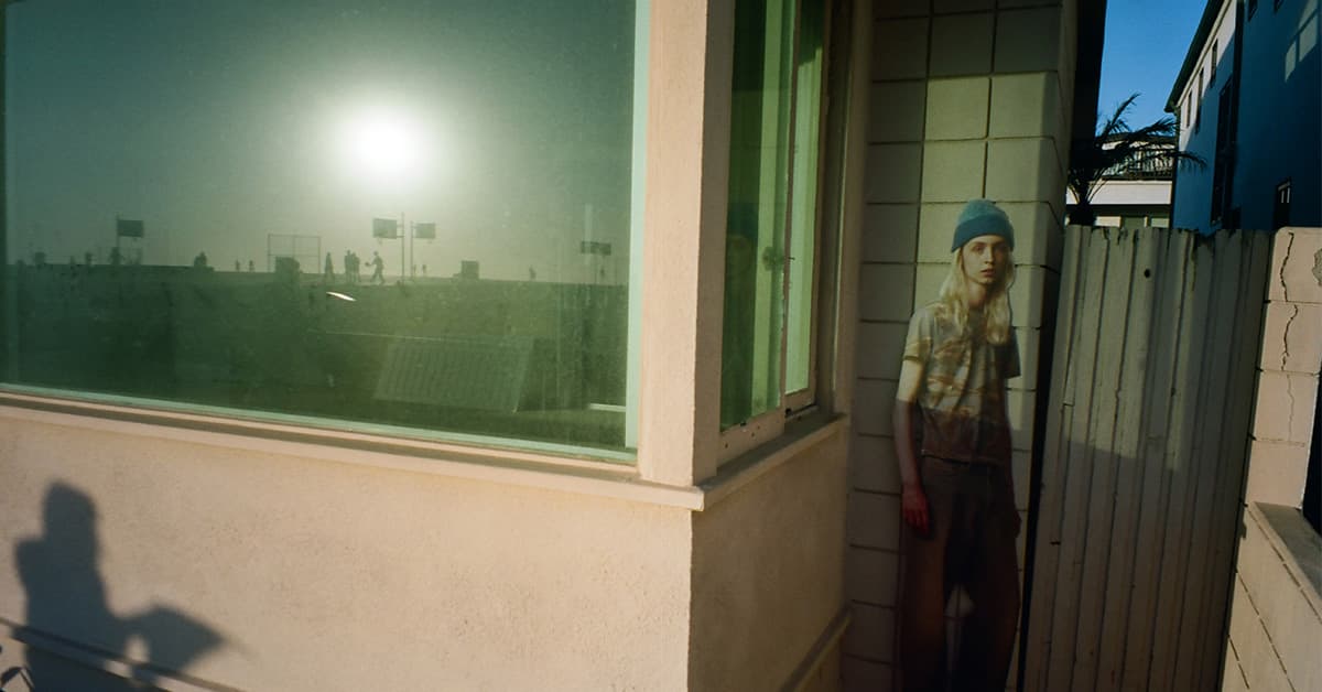A person with long blonde hair wearing a blue beanie and t-shirt standing near a building exterior with large green-tinted windows