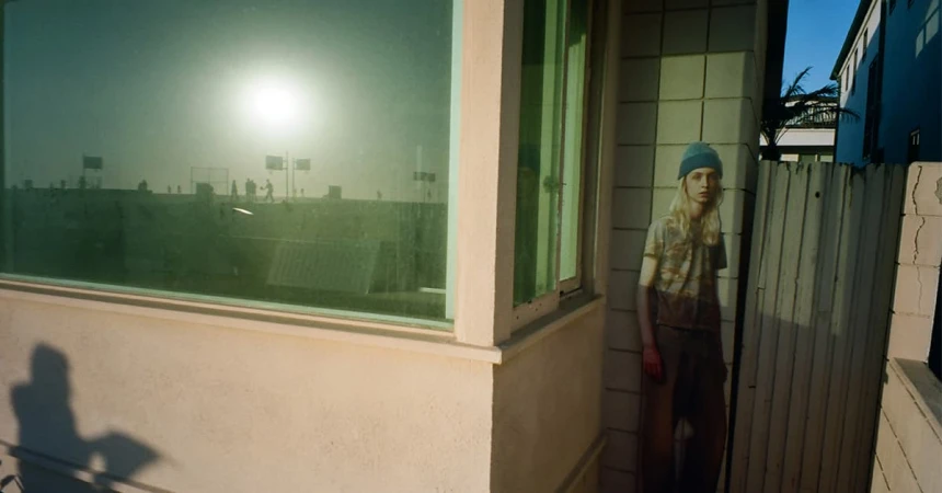 A person with long blonde hair wearing a blue beanie and t-shirt standing near a building exterior with large green-tinted windows