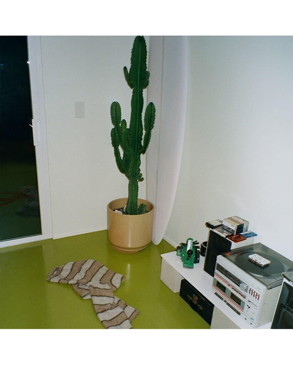 A tall potted cactus plant in a corner next to a white surfboard and a stereo system on a low white table.