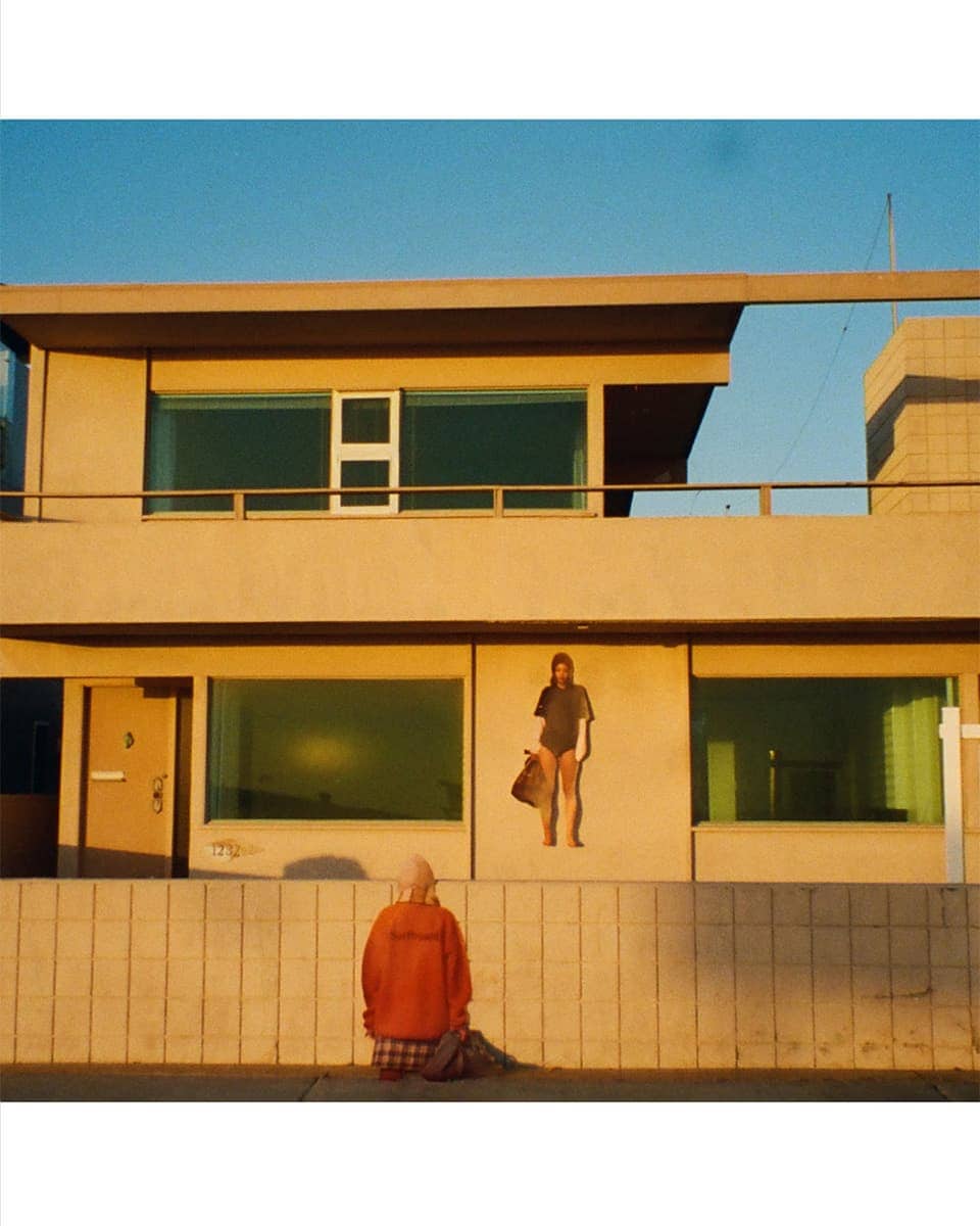 A person in an orange jacket standing before a modern multi-story stucco building under a clear blue sky.