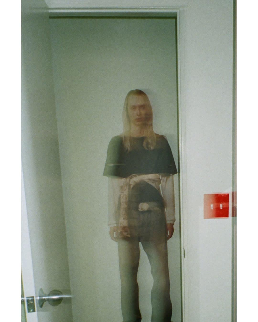 Long-haired person standing in a doorway wearing layered tops and jeans in an editorial portrait