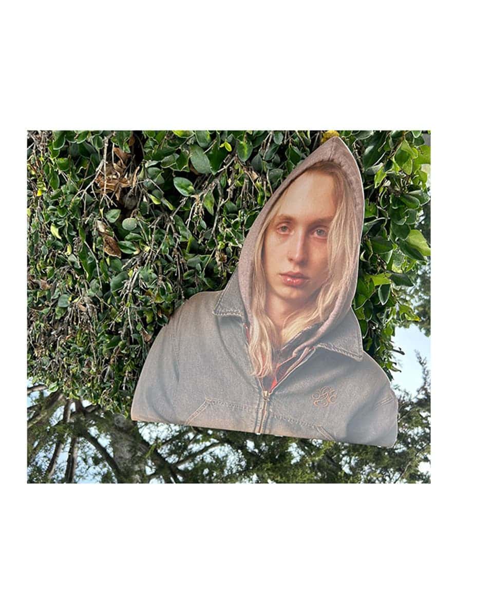 Portrait cut-out of a long-haired person in a hooded jacket mounted against green foliage and tree branches.