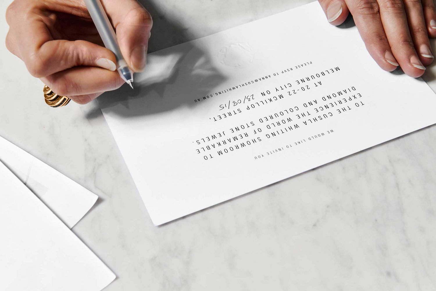 Close-up of hands writing on a printed white invitation card resting on a white marble surface.
