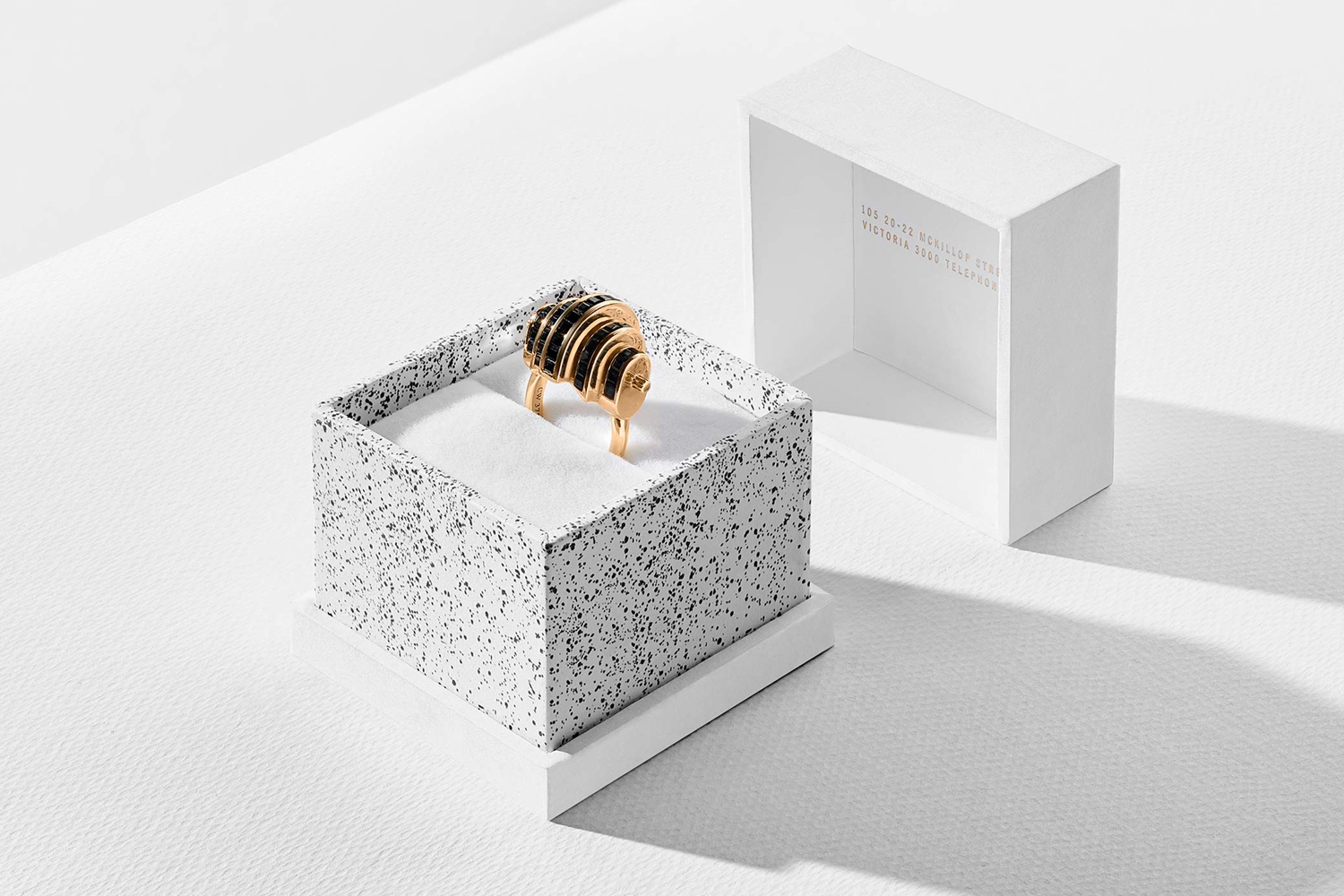 Gold ring with black details resting inside a speckled white box with a matching open lid on a white surface.