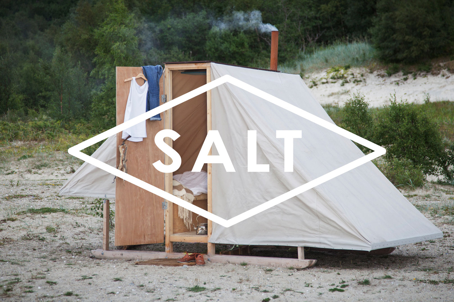 A small wooden cabin structure with a canvas cover set on a sandy beach, featuring a chimney with smoke.