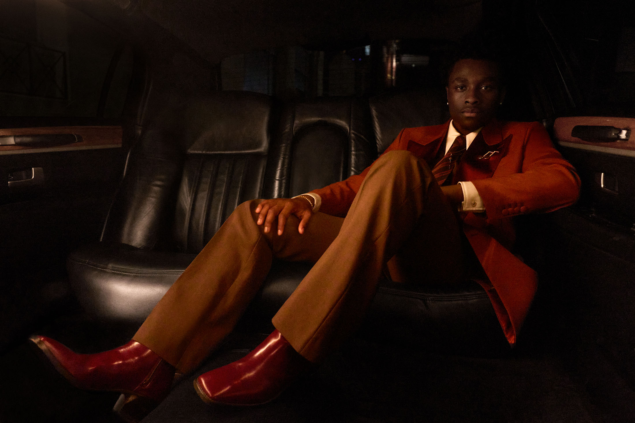 A young Black man seated inside a luxury car wearing a rust red blazer, brown trousers, and red leather boots.